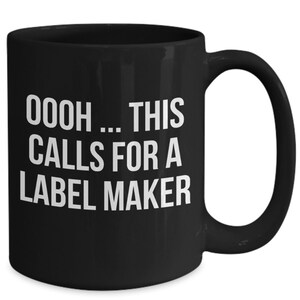 May include: Black ceramic coffee mug with white text that reads "OOOH... THIS CALLS FOR A LABEL MAKER".