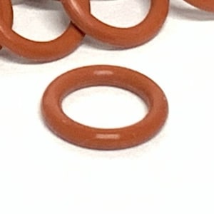 以下が含まれることがあります： 白い背景に散らばる茶色のゴム製Oリングのコレクション。Oリングは円形で、厚さが均一です。色は暖かく、アースカラーの茶色です。画像は明るく、リングの質感と形状を強調しています。
