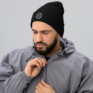 May include: Black beanie hat with a gray emblem. The hat has a ribbed texture and a turned-up brim. The emblem is a gray diamond shape within a gray circle. The person is wearing a gray jacket.