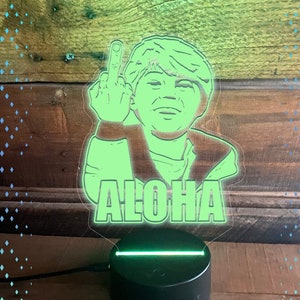 May include: An edge-lit night light featuring a laser-cut acrylic panel with a graphic of a person giving the middle finger and the text ALOHA. The panel has a 70mm bottom tab and sits on a black circular base. Includes .svg file and Glowforge text.