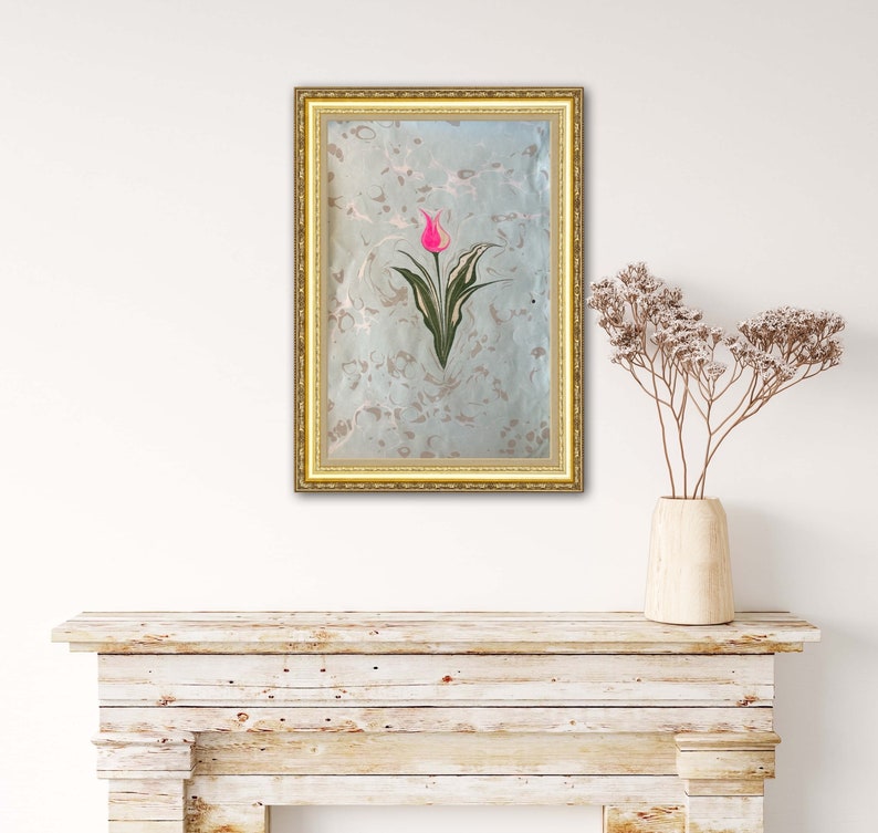 May include: A framed artwork featuring a pink tulip with green leaves on a light blue background with a marbled pattern. The frame is gold.