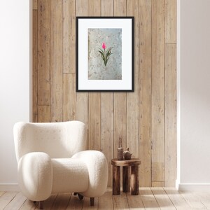 May include: A framed print of a pink tulip with green leaves on a white background. The print is hanging on a wood wall with a white armchair and a wooden side table in front of it.