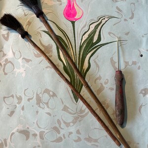 May include: Two wooden-handled brushes with black bristles and a small, thin, red-handled tool with a sharp point, all lying on a light blue and white marbled surface with a green and white tulip design.