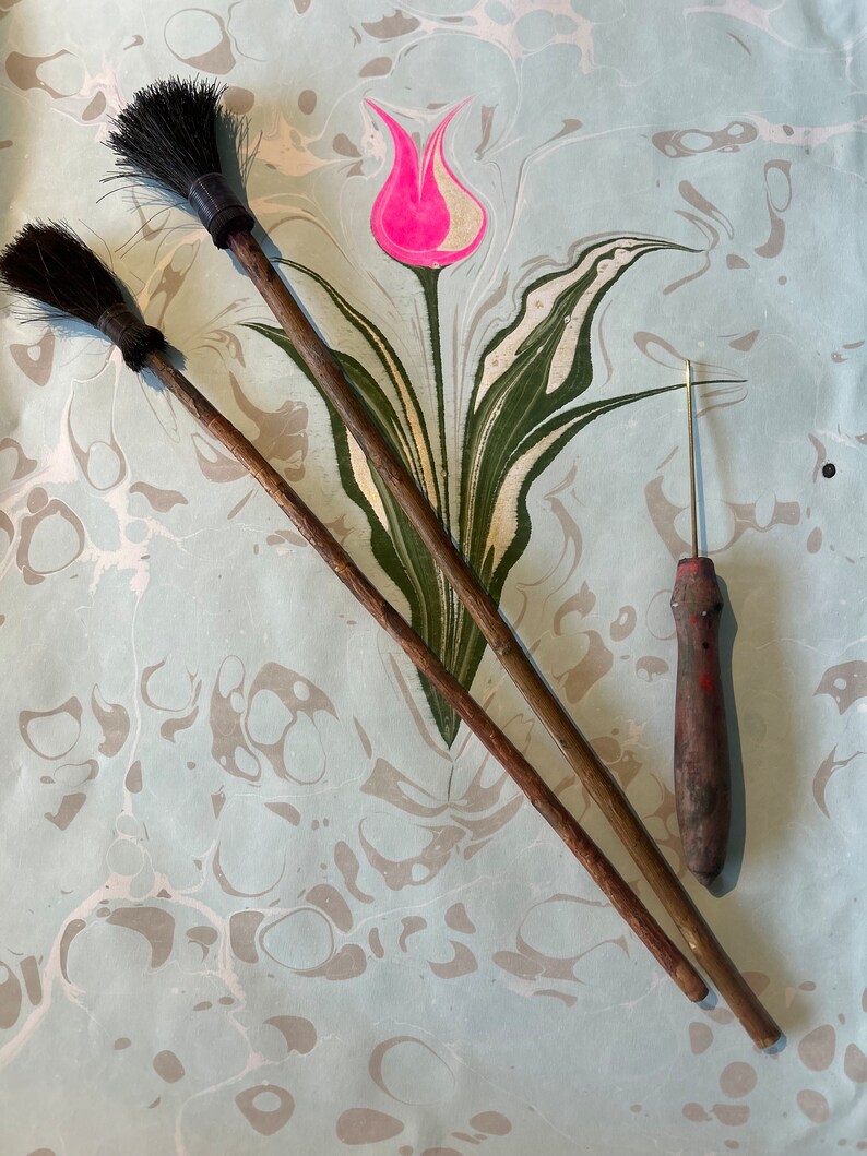 May include: Two wooden-handled brushes with black bristles and a small, thin, red-handled tool with a sharp point, all lying on a light blue and white marbled surface with a green and white tulip design.