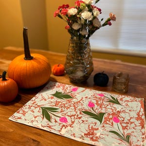 May include: A light blue and brown marbled placemat with pink flowers and green leaves. The placemat is on a wooden table with two pumpkins and a vase of flowers.