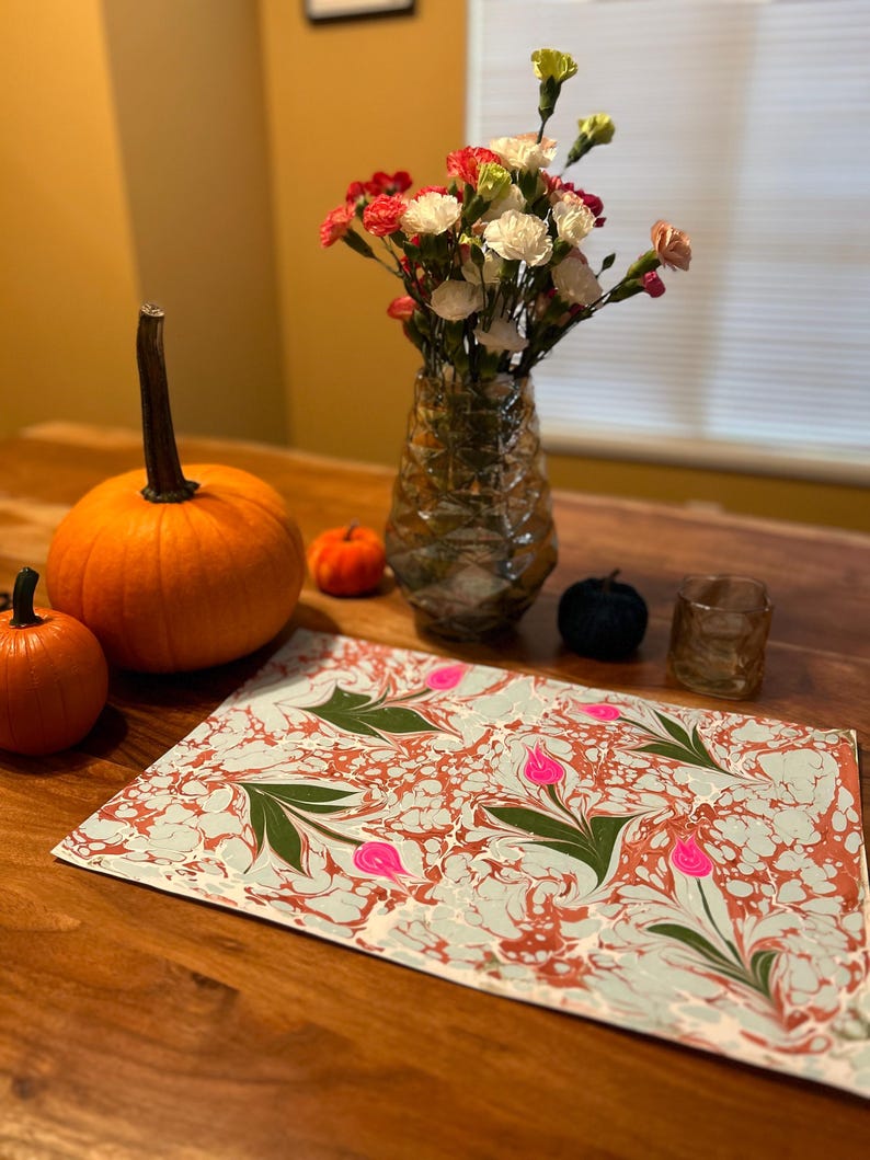 May include: A light blue and brown marbled placemat with pink flowers and green leaves. The placemat is on a wooden table with two pumpkins and a vase of flowers.