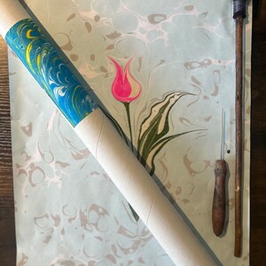 May include: A rolled up piece of marbled paper with a blue, green, and yellow design. The paper is on a wooden surface with a pink tulip design. There is a wooden tool with a brush on one end and a needle on the other end.