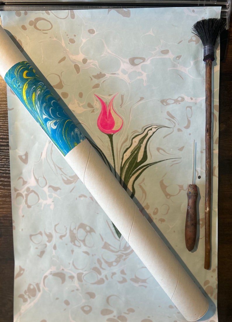 May include: A rolled up piece of marbled paper with a blue, green, and yellow design. The paper is on a wooden surface with a pink tulip design. There is a wooden tool with a brush on one end and a needle on the other end.