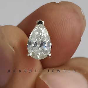 May include: A pear-shaped diamond gemstone. The diamond is held between two fingers and appears to be set in a small, silver or white metal setting. The diamond has a clear, sparkling appearance. The brand name 'BAARBIJ JEWELS' is visible in the image.