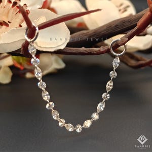 Può includere: Una catena d'argento con gemme chiare e scintillanti, che collega due cerchi d'argento. Il gioiello è presentato su uno sfondo scuro con un ramo e fiori bianchi. Il marchio "BAARBIJ JEWELS" è visibile.