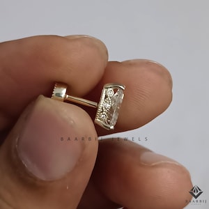 K&ouml;nnte beinhalten: Nahaufnahme eines goldfarbenen Ohrrings mit einem rechteckigen, klaren Edelstein. Der Ohrring hat ein Schraubverschluss-Design. Der Text "BAARBI JEWELS" ist auf dem Bild aufgedruckt.