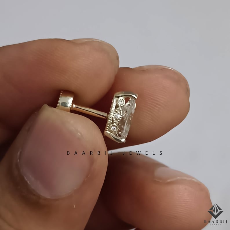 K&ouml;nnte beinhalten: Nahaufnahme eines goldfarbenen Ohrrings mit einem rechteckigen, klaren Edelstein. Der Ohrring hat ein Schraubverschluss-Design. Der Text "BAARBI JEWELS" ist auf dem Bild aufgedruckt.