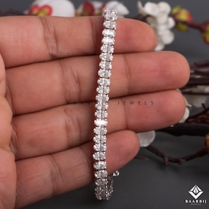 May include: A silver bracelet with a series of oval-cut, clear gemstones. The bracelet is held in a hand, with the brand name "BAARBIJ JEWELS" visible. The gemstones are set closely together, creating a continuous line of sparkle.