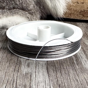 May include: A spool of dark gray wire sits on a weathered wooden surface. The wire is neatly wound around a white plastic spool. A strand of wire extends from the spool, suggesting its use in crafts or jewelry making.