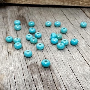 May include: A collection of turquoise-colored beads scattered on a weathered wooden surface. Each bead is round with a central hole, and the beads vary slightly in size. The wood has a natural, aged appearance with visible grain and cracks.