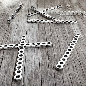 May include: Silver-toned metal jewelry components, including a cross shape, are arranged on a weathered wooden surface. The components have a series of circular cutouts. The background includes a fur-like texture and a metal-edged wooden object.