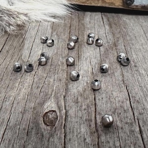 May include: Scattered on a weathered wooden surface are numerous small, metallic silver beads. The beads are round with a small hole. A section of a wooden box and a patch of fur are in the background.