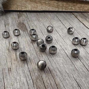 May include: A collection of small, metallic beads scattered on a weathered wooden surface. The beads are a dark silver color and have a rounded shape with a hole through the center. The wooden surface has a natural, rustic appearance.