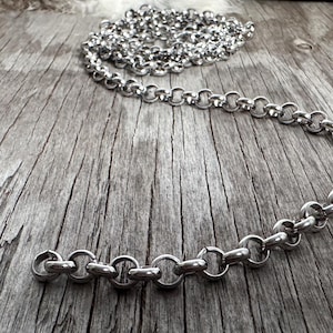 May include: A silver-colored chain necklace with large, round links is displayed on a weathered wooden surface. The necklace is partially coiled and partially extended, showcasing its design. The background includes a fur-like texture and a wooden element.