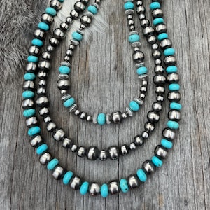 May include: Three-strand necklace featuring silver beads and turquoise-colored stones. The necklace is displayed on a weathered wooden surface, with a fur-like texture in the background. The necklace has a silver chain clasp.