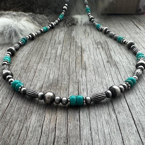 May include: A silver beaded necklace with turquoise accents. The necklace features a mix of round and cylindrical silver beads, interspersed with vibrant turquoise beads. The necklace is displayed on a weathered wooden surface.