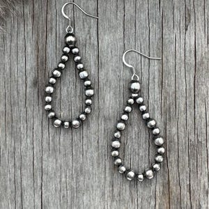 May include: A pair of silver teardrop earrings. Each earring features a series of small, round beads forming a teardrop shape. The earrings hang from a simple hook. The background is a weathered wooden surface.