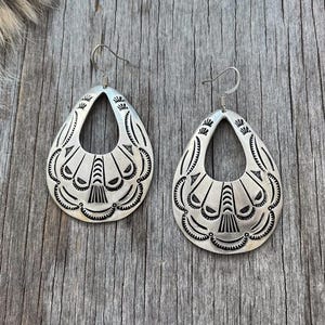 May include: Pair of silver-toned teardrop earrings with intricate stamped designs. Each earring features a central open space and detailed patterns along the edges, showcasing a Southwestern or Native American aesthetic.