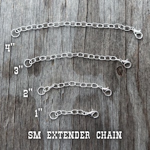May include: Four silver-colored SM extender chains of varying lengths: 1 inch, 2 inches, 3 inches, and 4 inches. Each chain has a lobster clasp and a jump ring. The chains are displayed on a weathered wooden surface.
