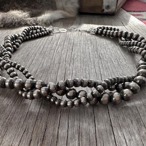 May include: A multi-strand silver beaded necklace with a secure clasp. The necklace features various sizes of round, dark silver beads, creating a layered effect. The jewelry is displayed on a rustic wooden surface.