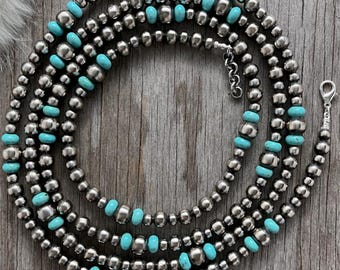 May include: A silver beaded necklace with turquoise accents. The necklace features multiple strands of small, round silver beads interspersed with vibrant turquoise beads. The necklace is displayed on a wooden surface.