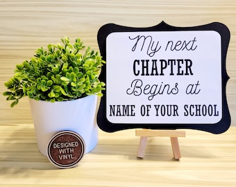 May include: Black and white sign with the text "My next CHAPTER Begins at NAME OF YOUR SCHOOL". The sign is on a small wooden easel. A small potted plant is in the foreground.