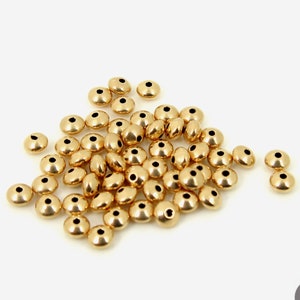 May include: A pile of gold-colored metal beads. The beads are round with a flat top and bottom. They are about 4 mm in diameter.