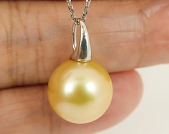 May include: A golden pearl pendant necklace. The large, round pearl has a lustrous, warm gold color. The pendant is attached to a delicate silver chain. The pendant's bail is a curved, silver-toned metal.