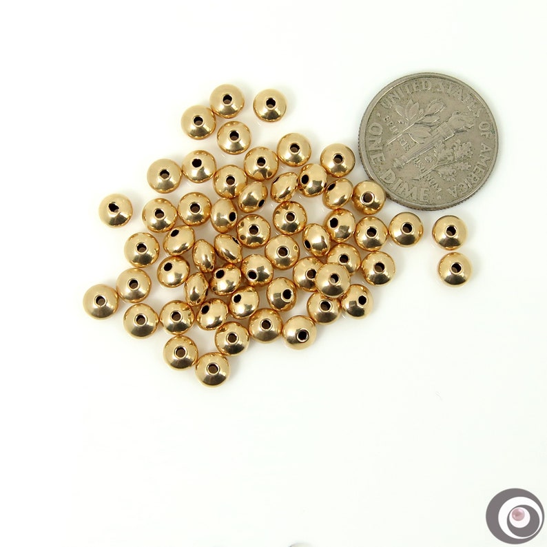 May include: A pile of gold-colored metal beads, some of which are slightly flattened, are scattered on a white surface. A US dime is included for scale.