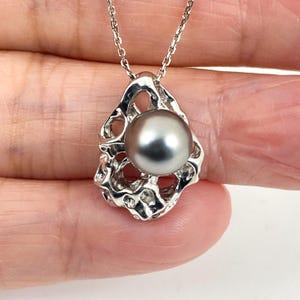 May include: A silver necklace featuring a gray pearl pendant. The pendant has an open, organic design surrounding the pearl. The chain is delicate and silver. The necklace is displayed against a neutral background.
