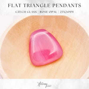 May include: A pink triangle-shaped pendant made of Czech glass. The pendant is 23x24mm and has a rose opal finish. The pendant is sitting on a wooden surface.