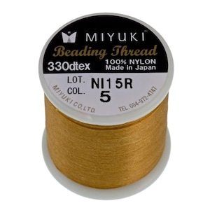 May include: A spool of golden-brown Miyuki beading thread made of 100% nylon. The white label on top features black text including the brand name, 330dtex, lot number NI15R, and color number 5. The thread is wound tightly around the cylindrical spool.