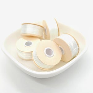 May include: A collection of white thread spools on a white ceramic dish. The spools are made of a light beige cardboard and are filled with a translucent, white thread. The spools are arranged in a slightly overlapping pattern.