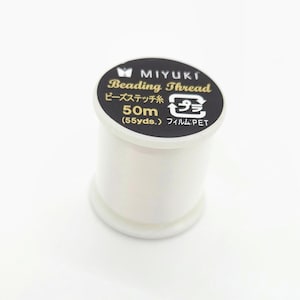 Op de afbeelding: Een witte spoel MIYUKI Beading Thread. Het zwarte etiket toont de merknaam, het type draad en de lengte van 50 m (55 yds). De plastic spoel is ontworpen voor kralenwerk en knutselprojecten.