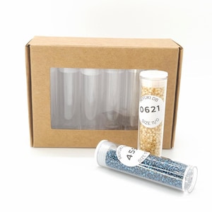 May include: A cardboard box with a window displaying several clear plastic tubes. Two tubes of beads are in front of the box. One tube contains gold beads, and the other contains blue beads. The gold beads tube has a label that reads "MIYUKI DB 0621 SIZE 11/0".