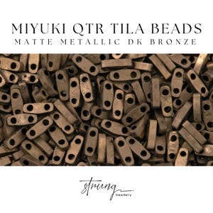 May include: Close-up of matte metallic DK bronze Miyuki QTR Tila beads. The beads are rectangular with two holes and a dark bronze color. The text "MIYUKI QTR TILA BEADS" and "MATTE METALLIC DK BRONZE" are at the top. The logo for "strung beadery" is at the bottom.