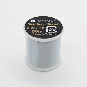 May include: A spool of Miyuki Beading Thread in a light gray color. The label is black with gold lettering and includes the text "Beading Thread", "50m (55 yds.)", and Japanese text. The spool is made of plastic.