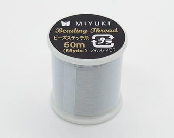 May include: A spool of Miyuki Beading Thread in a light gray color. The label is black with gold lettering and includes the text "Beading Thread", "50m (55 yds.)", and Japanese text. The spool is made of plastic.