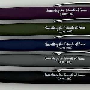 May include: A collection of five pens in various colors: purple, olive green, navy blue, gray, and black. Each pen has a silver tip and clip, with the text "Searching for Friends of Peace (LUKE 10:6)" printed on the barrel.