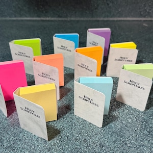 May include: A collection of small, book-shaped sticky note pads with beige covers printed with the text Holy Scriptures. Each pad features a different colored interior paper, including neon pink, yellow, orange, blue, green, and purple.