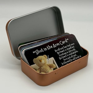 May include: A copper-colored metal tin containing a stack of rectangular cards. The front card features a tan teddy bear, a small syringe, and the text "Shot in the Arm Card" followed by religious scripture about trusting in Jehovah.