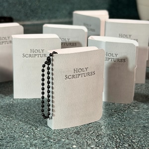 May include: A collection of small, white, rectangular books with the text HOLY SCRIPTURES embossed on the front cover. Each book features a black ball chain attached to the spine, allowing it to function as a keychain or hanging ornament.