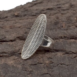 Könnte beinhalten: Ein silberner Ring mit einer länglichen ovalen Oberfläche mit einem strukturierten Fischgrätenmuster. Der Ring ist auf einer dunklen, strukturierten Holzoberfläche platziert, die das komplizierte Design und den metallischen Glanz hervorhebt.