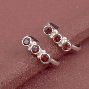 Könnte beinhalten: Ein Paar silberne Zehenringe, jeweils mit drei runden, tiefroten Edelsteinen. Die Ringe sind auf einer braunen Oberfläche platziert, die das Design und den Farbkontrast des Schmucks hervorhebt. Die Edelsteine sind gleichmäßig verteilt.