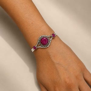 Peut inclure: Un bracelet manchette en argent orné d'une grande pierre précieuse magenta ronde et facettée au centre, flanquée de deux pierres magenta rondes et facettées plus petites. La monture en argent présente un motif de perles métalliques.